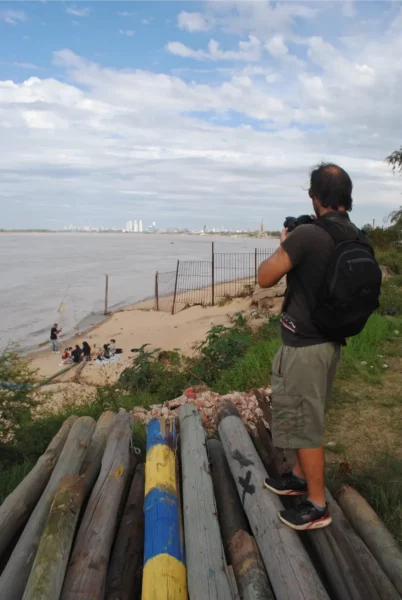 A photographer captures the riverbank scene at Rosario, Santa Fe, Argentina, framed by wooden logs and a cloudy sky.