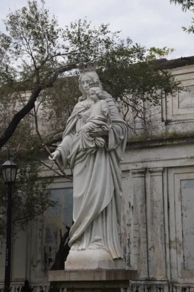 White statue of a saint holding a child, surrounded by trees, in a historic area of Rosario, Santa Fe, Argentina.