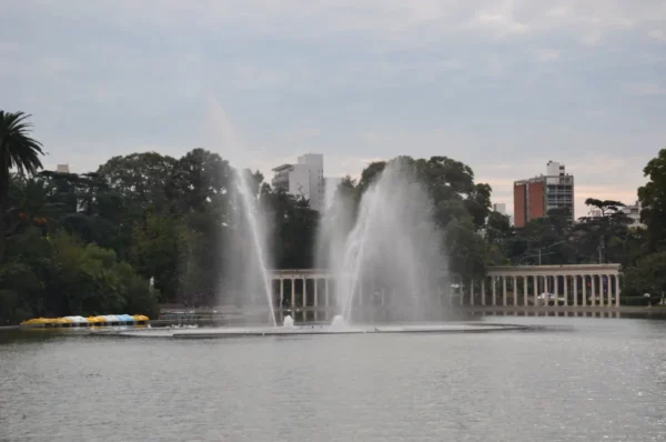 Fountain sprays create a striking scene at the lake in Rosario, Santa Fe, Argentina, surrounded by lush greenery and city skyline.