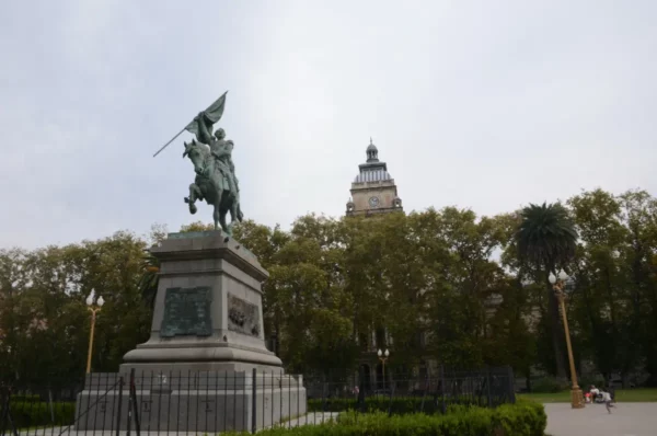 Equestrian statue in Rosario, Santa Fe, Argentina, with a historic building and lush trees in the background.