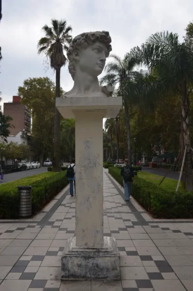 David's sculpture head on a pedestal, surrounded by palm trees and greenery along a tiled pathway in Rosario, Santa Fe, Argentina.
