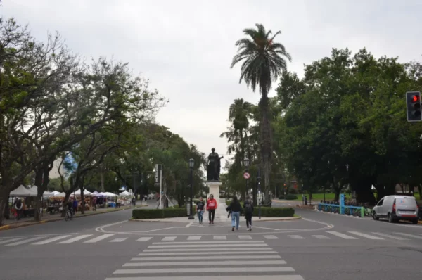 Statue at the center of a tree-lined avenue in Rosario, Santa Fe, Argentina, with pedestrians crossing streets under a gray sky.