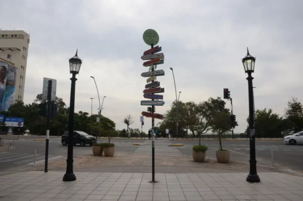 Colorful directional signs stand tall amid street lamps and greenery in Rosario, Santa Fe, Argentina against a cloudy sky.