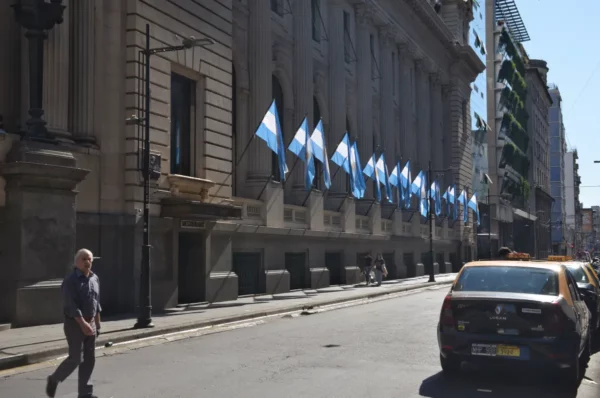 Blue and white flags adorn the building facade along a sunny street in Rosario, Santa Fe, Argentina, with a passing taxi.
