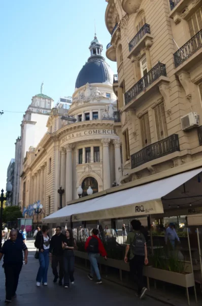 Elegant architecture lines the street in Rosario, with a prominent dome and bustling outdoor seating. Sunny weather enhances the scene.