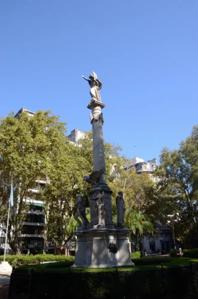 Statue atop a tall column surrounded by greenery, with modern buildings and a clear blue sky in Rosario, Santa Fe, Argentina.