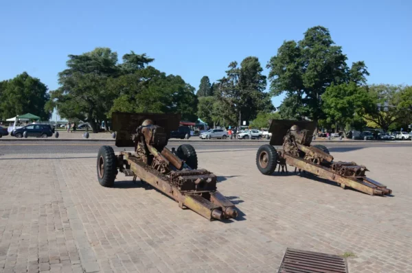 Rusty artillery pieces are displayed on a cobblestone plaza surrounded by trees in Rosario, Santa Fe, Argentina.