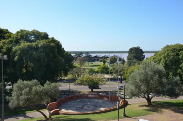 Vibrant green trees frame a scenic view of the Paraná River in Rosario, Santa Fe, showcasing parkland and blue skies.
