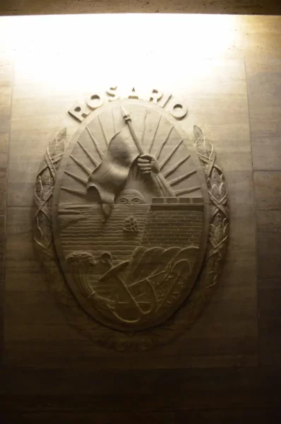 Rosario emblem featuring a stylized sunburst and a symbolic figure, illuminated against a textured wall in Santa Fe, Argentina.