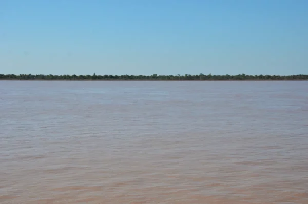 Calm waters of the Paraná River reflecting a clear sky near Rosario, Santa Fe, Argentina, with distant greenery on the horizon.
