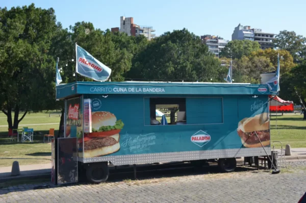 Colorful food cart with flags serving burgers, set against lush greenery in a park in Rosario, Santa Fe, Argentina.