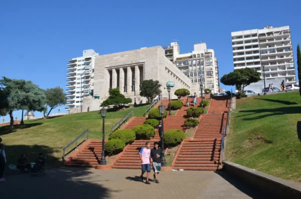 Stairs lead up to a grand building surrounded by greenery in Rosario, Santa Fe, Argentina, under a clear blue sky.