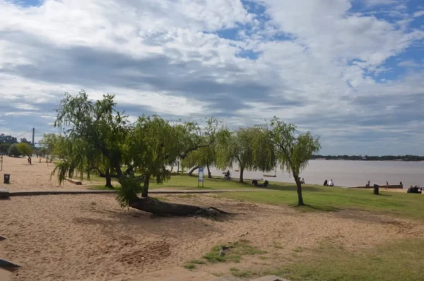 Sunny riverside park in Rosario, Santa Fe, Argentina, featuring green trees, sandy areas, and a view of the river under a cloudy sky.