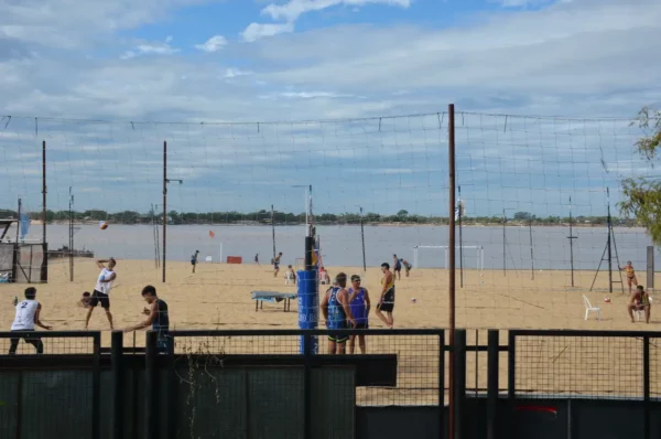 Beach volleyball games take place on the sandy shores of Rosario, Santa Fe, Argentina, with the river and cloudy sky in the background.