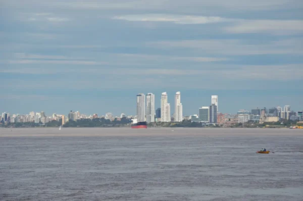 Modern skyline of Rosario, Santa Fe, Argentina, with high-rise buildings and a calm river in the foreground under a blue sky.