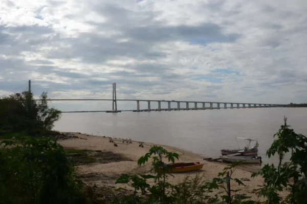Rosario's iconic bridge spans the river, framed by a sandy shore and scattered greenery, under a cloudy sky in Santa Fe, Argentina.