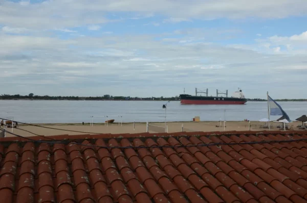 Large cargo ship sailing on the Paraná River, with terracotta rooftops and a cloudy sky in Rosario, Santa Fe, Argentina.