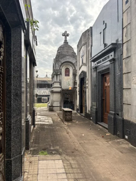 Quiet pathway in Rosario, Santa Fe, Argentina, flanked by mausoleums and a cross-topped structure under a cloudy sky.