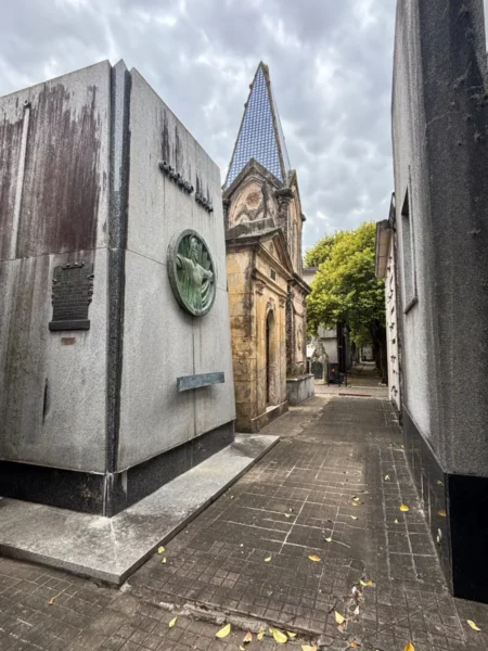 Cemetery pathway in Rosario, Santa Fe, Argentina, lined with unique mausoleums and trees under a cloudy sky.