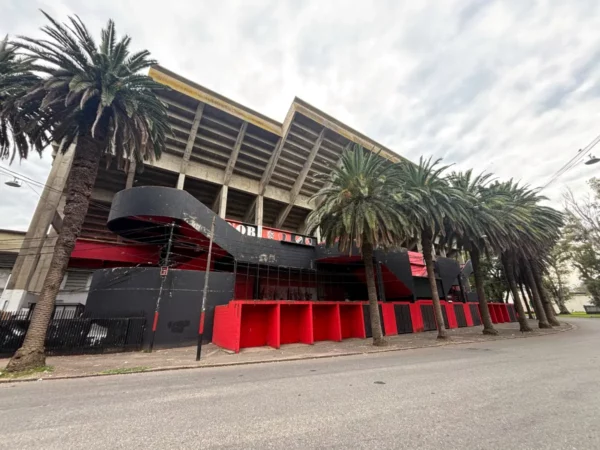 Modern stadium with red and black accents surrounded by tall palm trees in Rosario, Santa Fe, Argentina. Cloudy sky above.