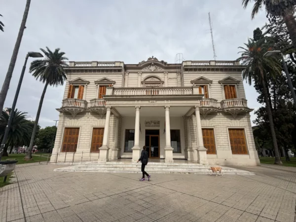 Elegant white mansion with detailed architecture, surrounded by palm trees in Rosario, Santa Fe, Argentina. A person and dog are visible in the foreground.