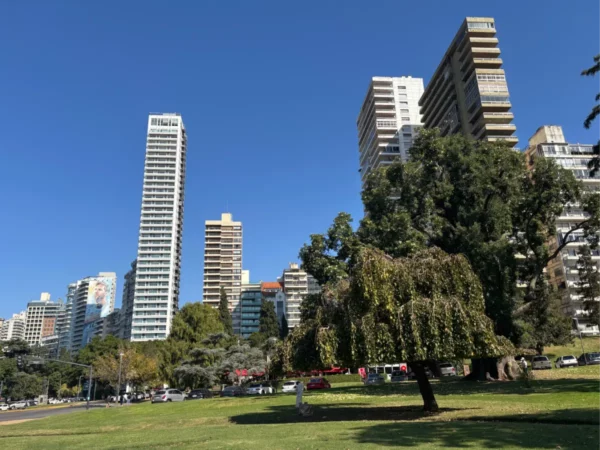 Tall skyscrapers rise against a clear blue sky in Rosario, Santa Fe, Argentina, surrounded by lush greenery and urban parkland.