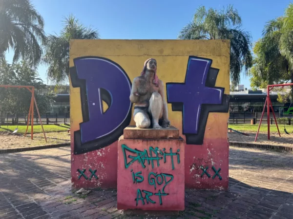 Colorful mural with graffiti and a statue in a park setting, featuring a bright yellow and pink background in Rosario, Santa Fe, Argentina.