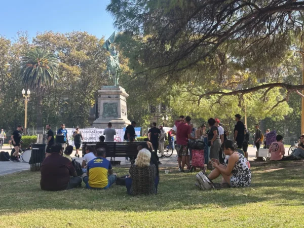 People relax on the grass near a statue in Rosario, Santa Fe, Argentina, surrounded by lush trees and a sunny atmosphere.