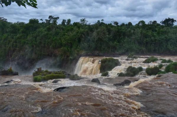 Rushing waters cascade over Saltos del Monday Falls, surrounded by lush greenery and a cloudy sky in Paraguay.