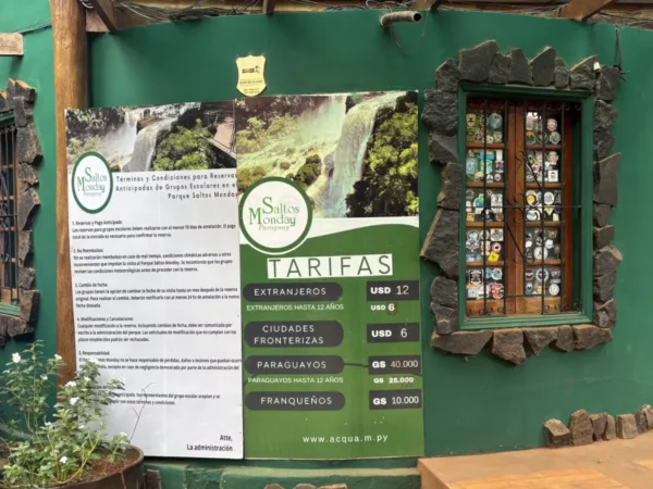 Informative sign displaying entrance fees and visitor information for Saltos del Monday Falls in Paraguay, with a vibrant green backdrop.