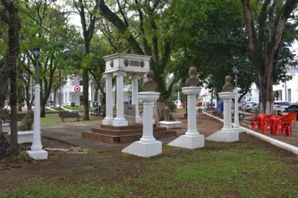 Busts and a white gazebo surrounded by lush trees in a green park in Villarica, Paraguay. Red chairs add a vibrant touch.