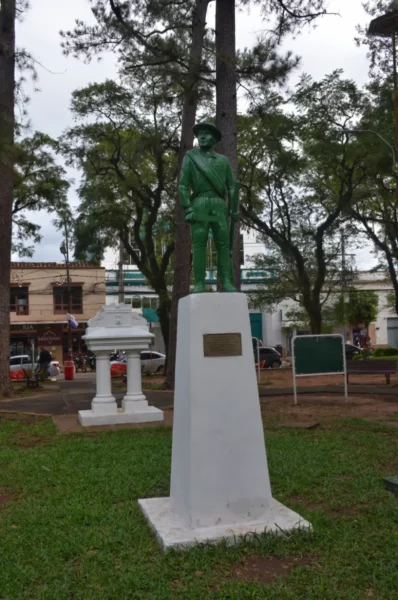 Green statue of a soldier atop a white pedestal surrounded by trees in Villarica, Paraguay, with a park-like setting.