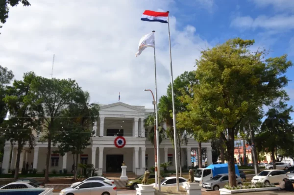 White colonial building adorned with flags, surrounded by trees and a clear blue sky in Villarica, Paraguay.