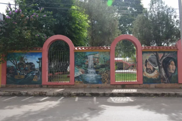 Colorful murals depicting local scenes adorn a pink wall, surrounded by lush greenery in Villarica, Paraguay.