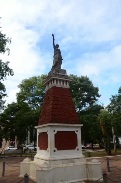 Statue atop a red pyramid monument, surrounded by green trees under a blue sky in Villarica, Paraguay.