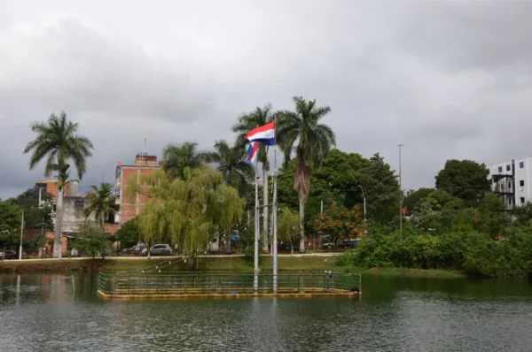 Paraguayan flags stand proudly near a serene pond surrounded by lush greenery and palm trees in Villarica, Paraguay.