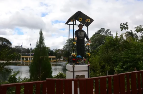 Statue surrounded by greenery and sunflowers near a tranquil body of water in Villarica, Paraguay. Cloudy sky above.