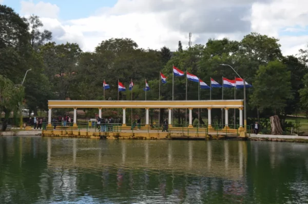 Vibrant flags wave over the serene waters of Villarica, Paraguay, with green trees framing the picturesque scene.