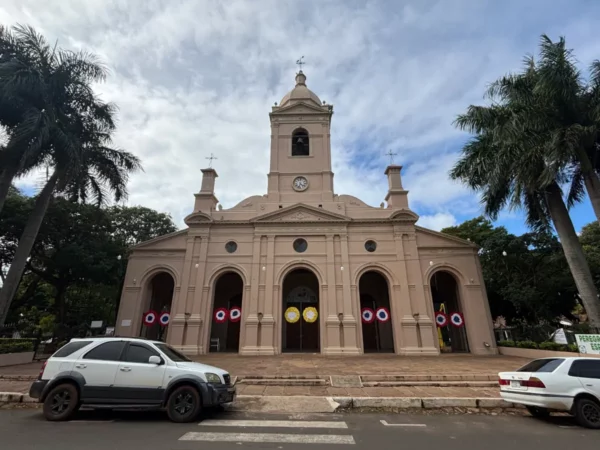 Pink church facade adorned with decorative wreaths and palm trees in Villarica, Paraguay, under a partly cloudy sky.