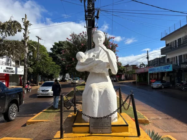 A white statue of a woman holding a baby contrasts against a blue sky in Villarica, Paraguay, surrounded by urban streets.