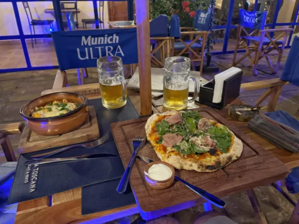 Delicious pizza topped with greens and ham, accompanied by a bowl of soup and two glasses of beer at a lively outdoor restaurant in Villarica, Paraguay.