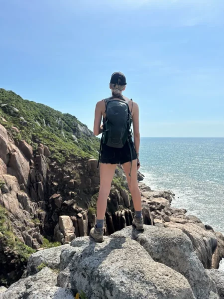 Hiker stands on rocky cliffs overlooking the ocean at Praia do Rosa, Santa Catarina, Brazil, with clear blue skies above.