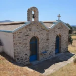 Charming stone chapel with blue doors and a bell tower, set in the hills of Karystos, Euboea, Greece. Sunlit landscape surrounds it.
