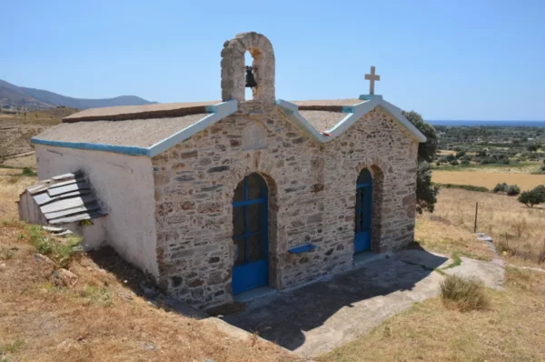 Charming stone chapel with blue doors and a bell tower, set in the hills of Karystos, Euboea, Greece. Sunlit landscape surrounds it.