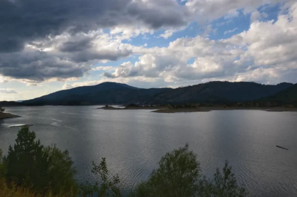 Serene Aoos Lake reflects the gray sky and surrounding hills near Ioannina, Greece, surrounded by lush greenery.