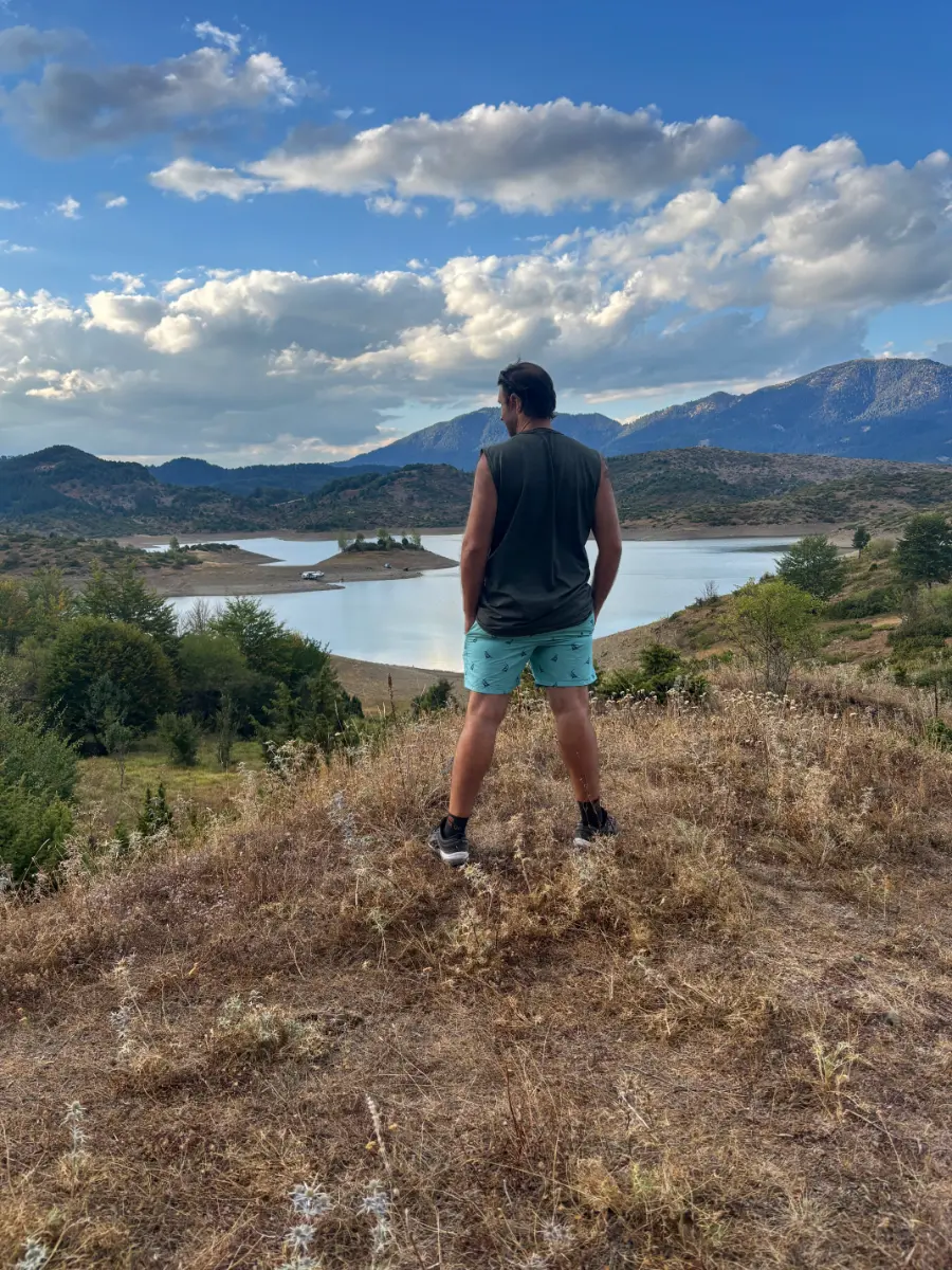 Man standing on a hillside overlooking Aoos Springs Lake, with mountains, clouds, and forested islets in northern Greece