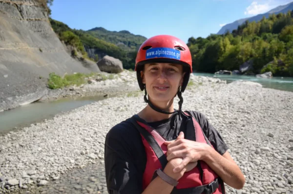 Rafting enthusiast in a red helmet stands on pebbly shore near the Arachthos River, surrounded by lush green hills in Ioannina, Greece.