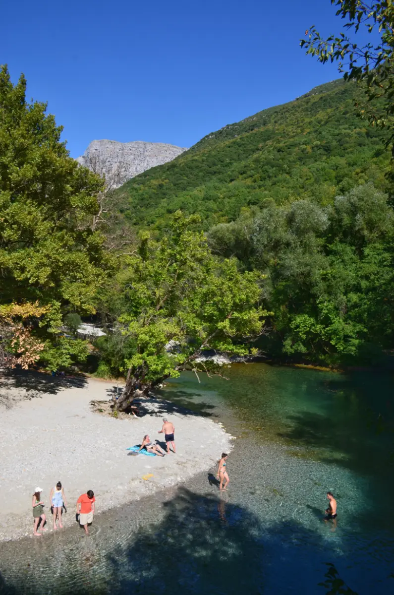 Pebble beach by the Aristi stone bridge on the Voidomatis River, Zagorochoria, with clear turquoise water, swimmers cooling off, and forested limestone slopes in summer.