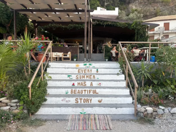 Charming steps lead up to a café at Chiliadou Beach, Euboea, Greece, adorned with a welcoming quote and lush greenery.