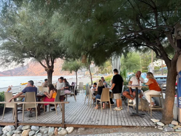 Chiliadou Beach in Euboea, Greece, features beachgoers dining under trees at outdoor tables, with a sandy shore in the background.